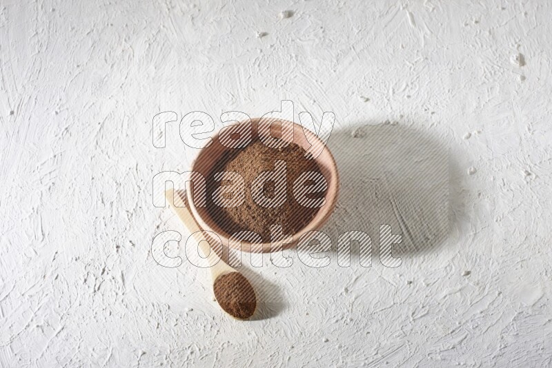 A wooden bowl and a wooden spoon full of cloves powder on a textured white flooring