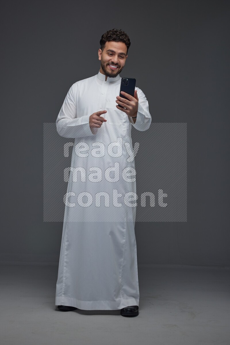 A Saudi man wearing Thobe and making a video call using his phone while standing and making different poses eye level on a gray background