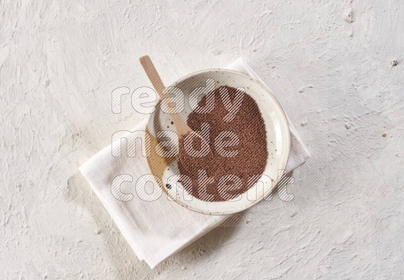 A multicolored pottery plate full of garden cress seeds with a wooden spoon full of the seeds on a napkin on a textured white flooring