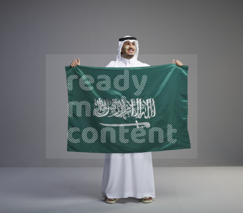 A Saudi man standing wearing thob and white shomag with face painting holding big Saudi flag on gray background