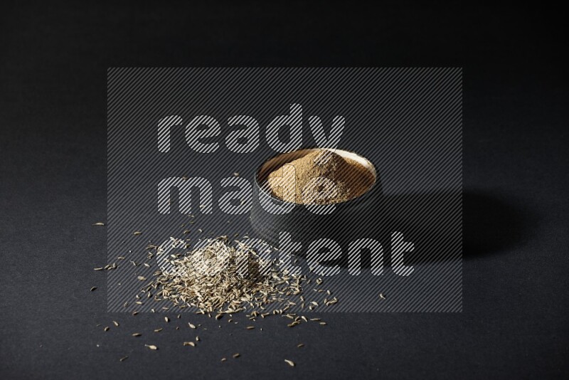 A black pottery bowl full of cumin powder and spreaded cumin seeds on a black flooring