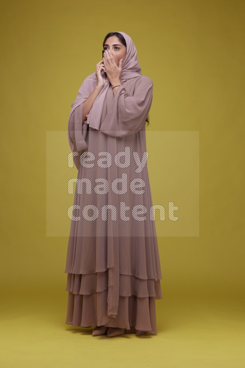 A woman having a call on a Yellow Background wearing Brown Abaya with Hijab