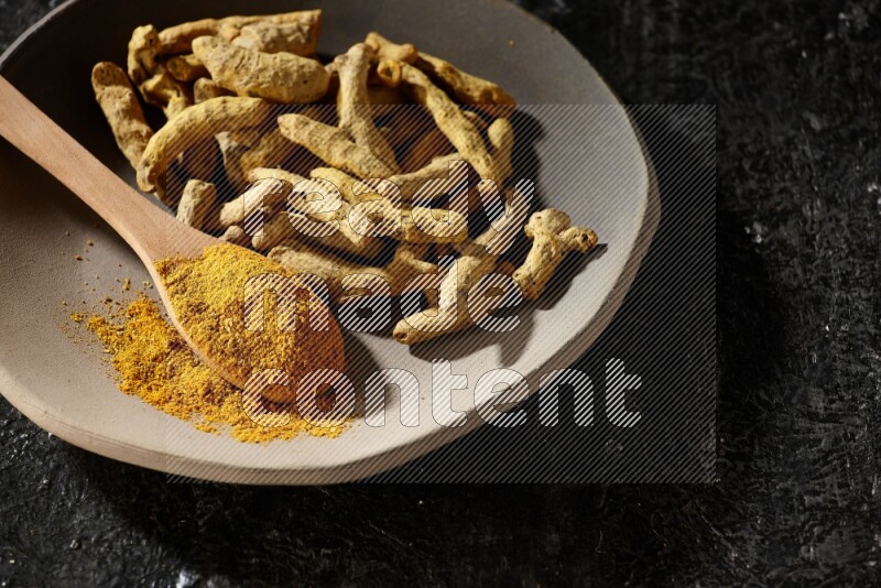 A plate filled with dried turmeric fingers and a wooden spoon full of turmeric powder on a textured black flooring