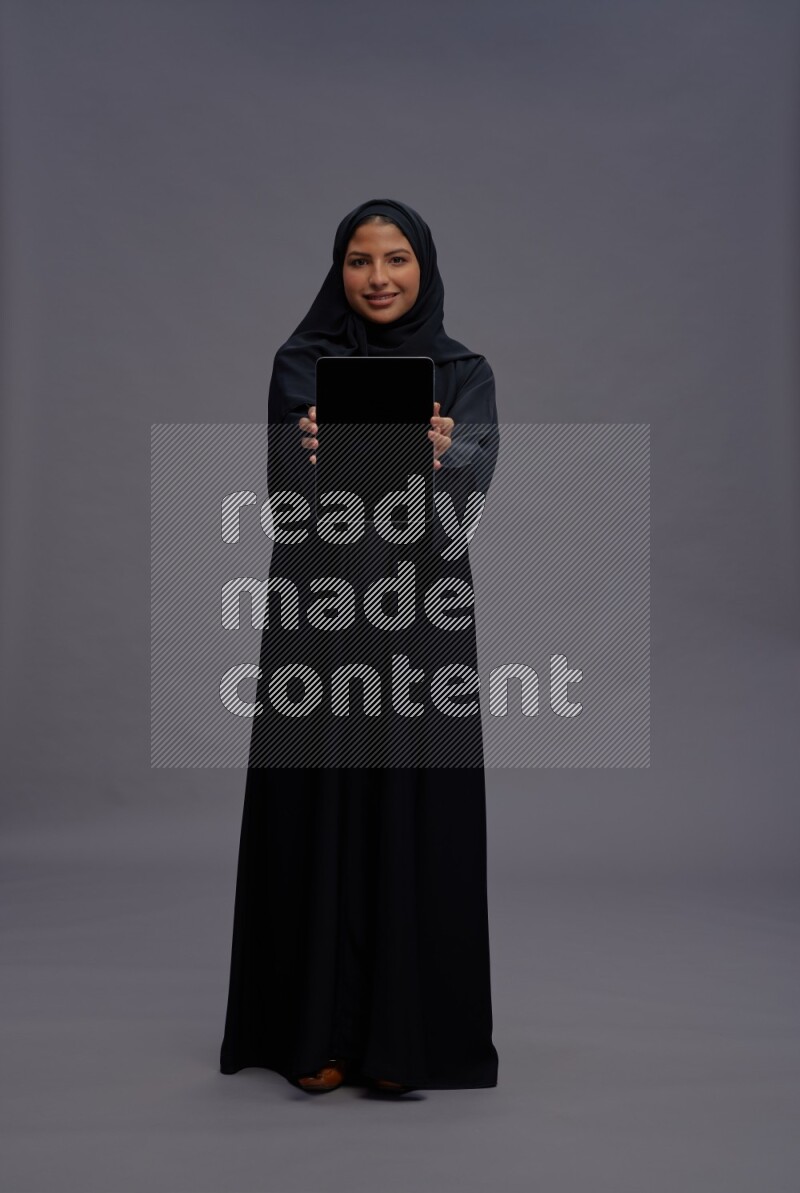 Saudi woman wearing Abaya standing showing tablet to camera on gray background