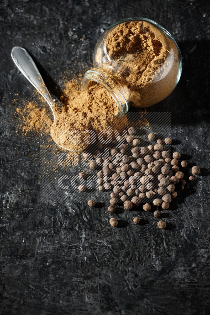 A flipped glass spice jar and metal spoon full of allspice powder and powder spilled out of it with whole balls on a textured black flooring