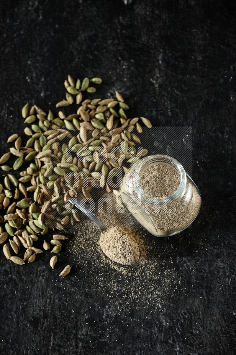 A glass spice jar and a metal spoon full of cardamom powder and cardamom seeds spreaded on textured black flooring