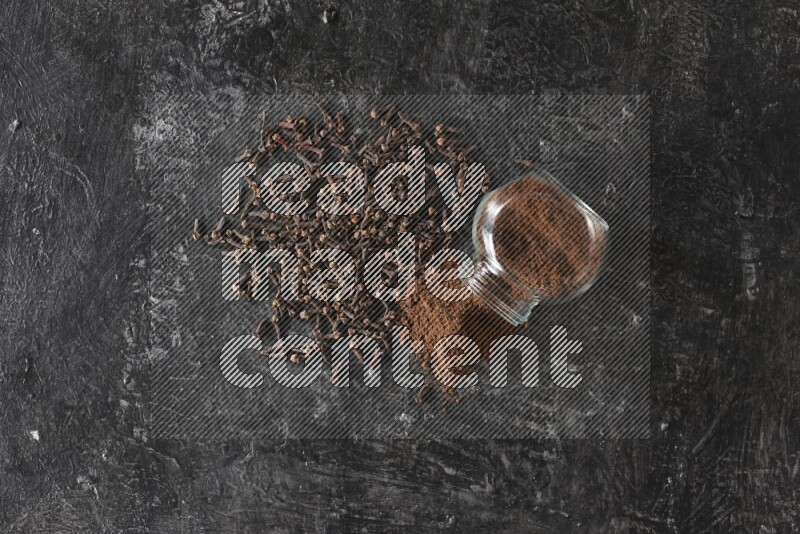 A flipped glass spice jar full of cloves powder and powder came out of it with cloves spread on textured black flooring