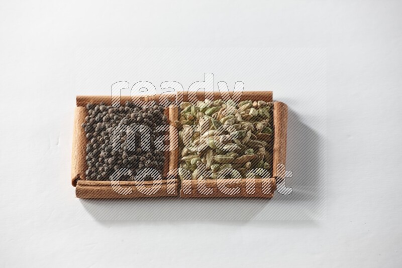 مربعان من أعواد القرفة ممتلئان بحبوب الهيل وحبوب الفلفل الأسود علي خلفية بيضاء