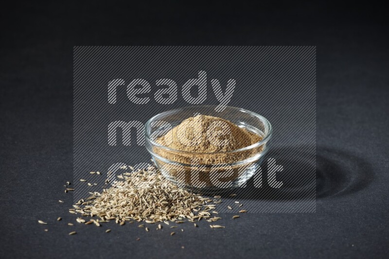 A glass bowl full of cumin powder with cumin seeds beside it on black flooring