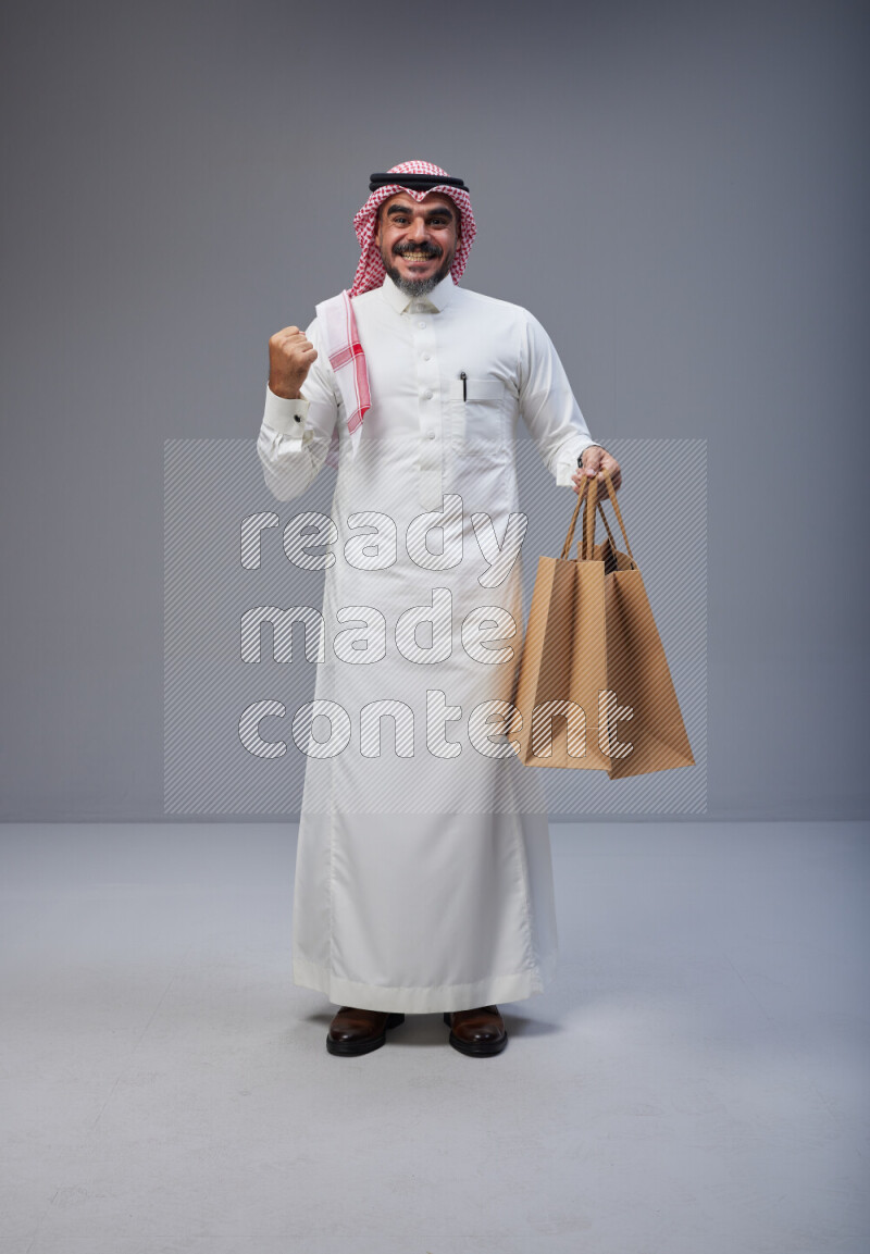 Saudi man Wearing Thob and red Shomag standing holding shopping bag on Gray background