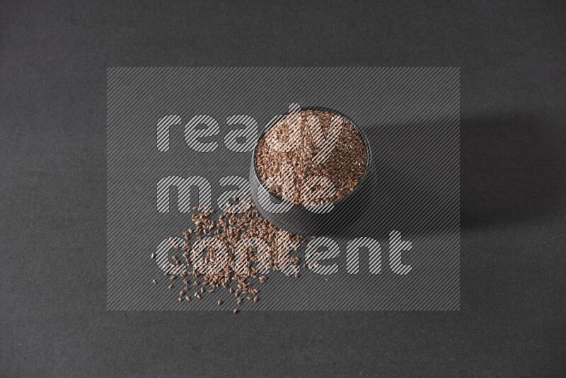 A black pottery bowl full of flaxseeds surrounded by the seeds on a black flooring