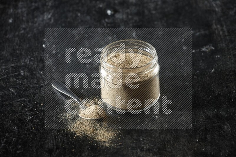 A glass jar and metal spoon full of cardamom powder on textured black flooring