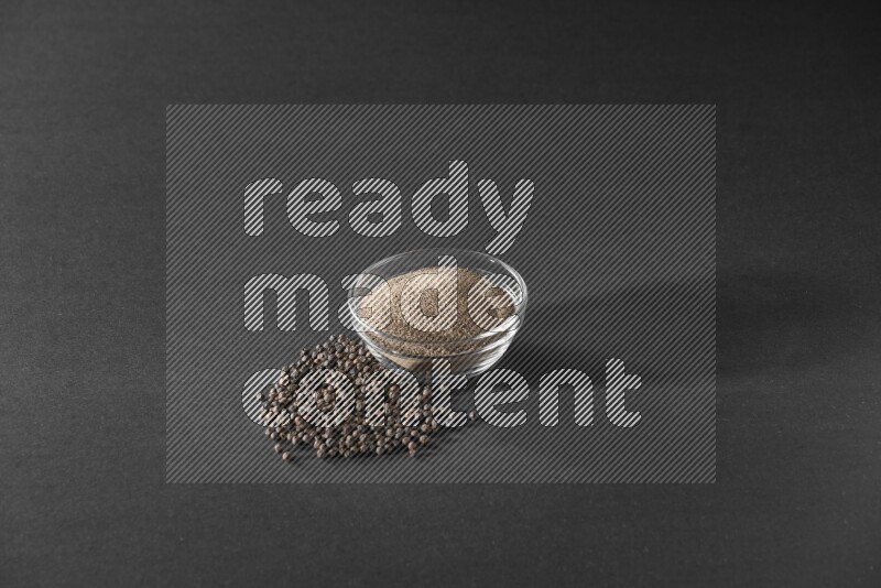 A glass bowl full of powder black pepper powder and black pepper beads on the floor on black flooring