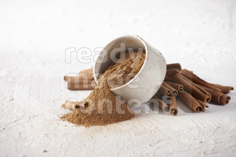 Ceramic beige bowl over filled with cinnamon powder and cinnamon sticks around the bowl on a textured white background in different angles