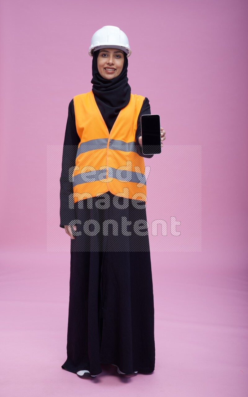Saudi woman wearing Abaya with engineer vest and helmet standing showing phone to camera on pink background