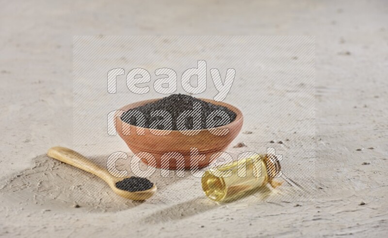 A wooden bowl and spoon full of black seeds with a bottle of black seeds oil on a textured white flooring