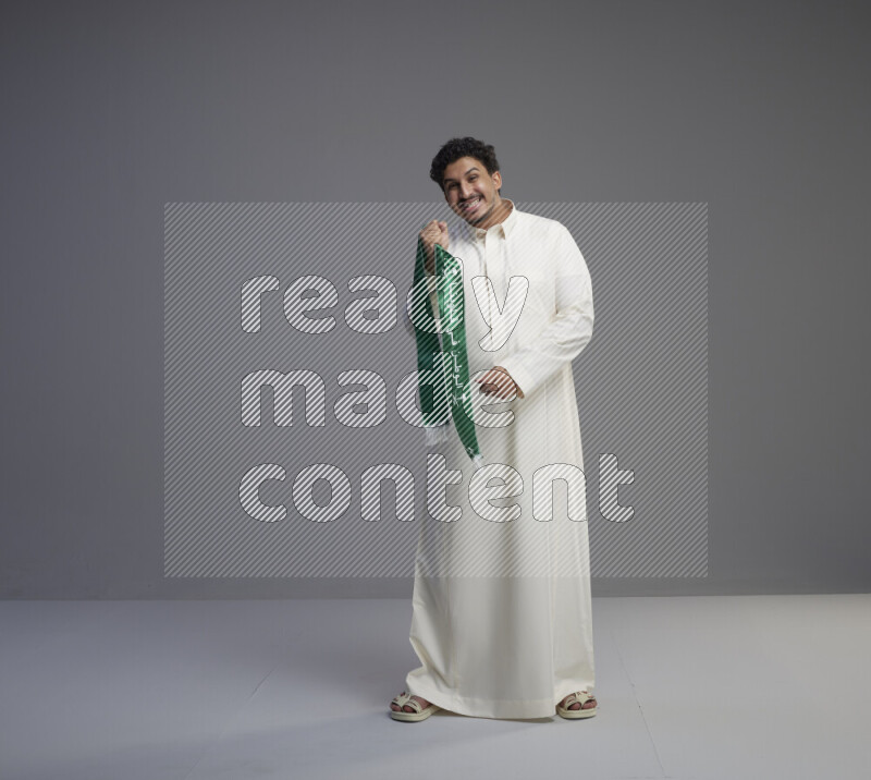 A Saudi man standing wearing thob holding Saudi flag scarf on gray background