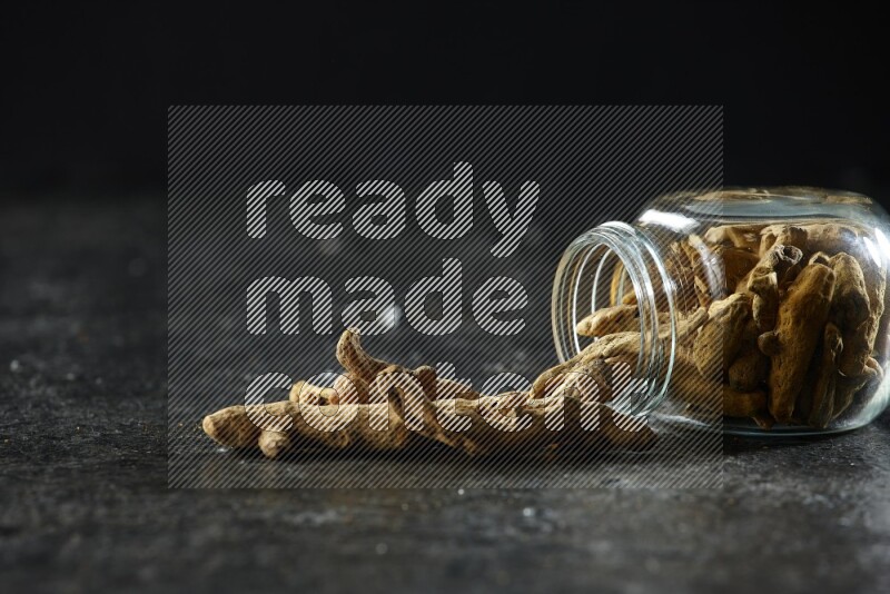 A flipped glass spice jar full of dried turmeric whole fingers and fingers fell out of it on textured black flooring