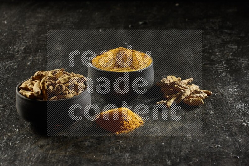 2 pottery black bowls, one full of turmeric powder and the other full of dried turmeric whole fingers on a textured black flooring