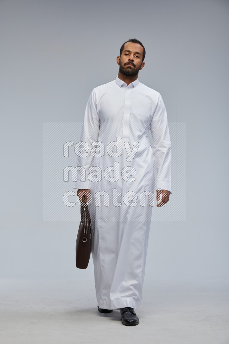 Saudi man wearing thob standing holding suitcase on gray background