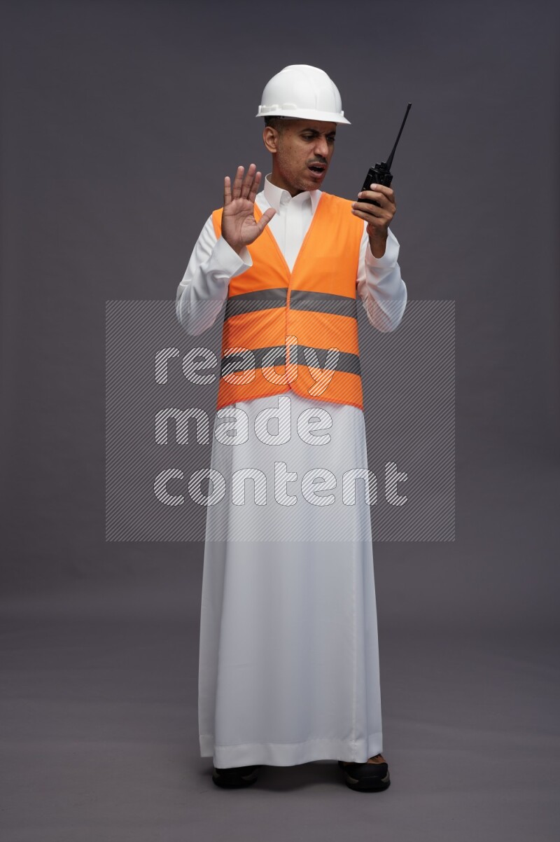 Saudi man wearing thob with engineer vest standing holding walkie-talkie on gray background