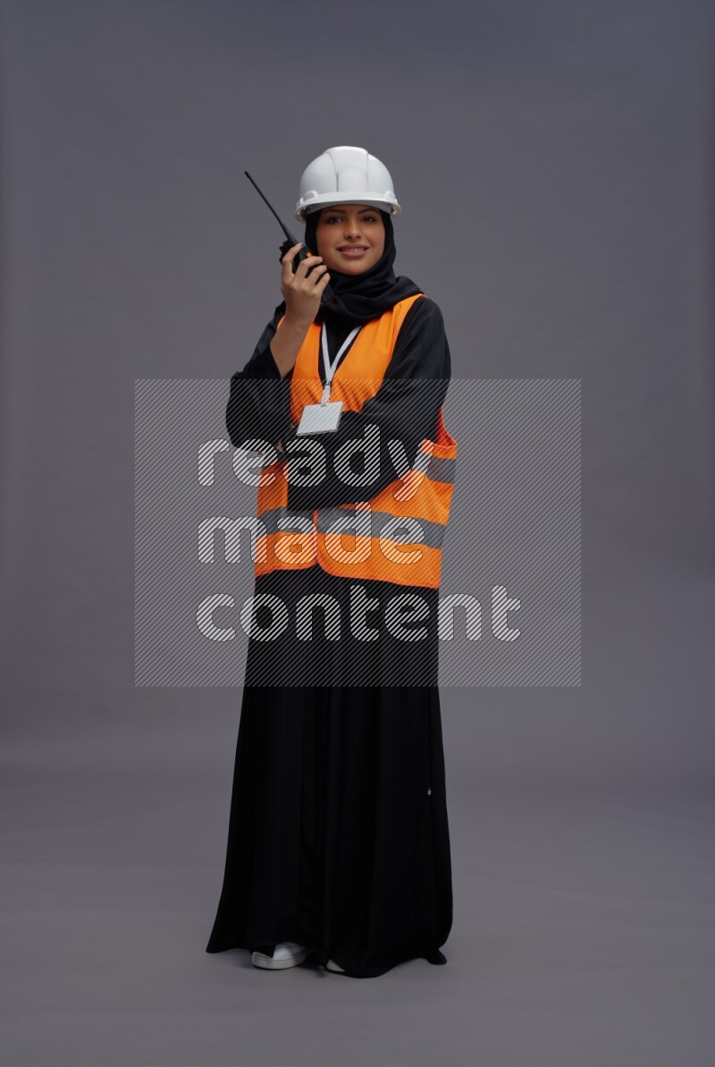Saudi woman wearing Abaya with engineer vest with neck strap employee badge standing holding walkie-talkie on gray background