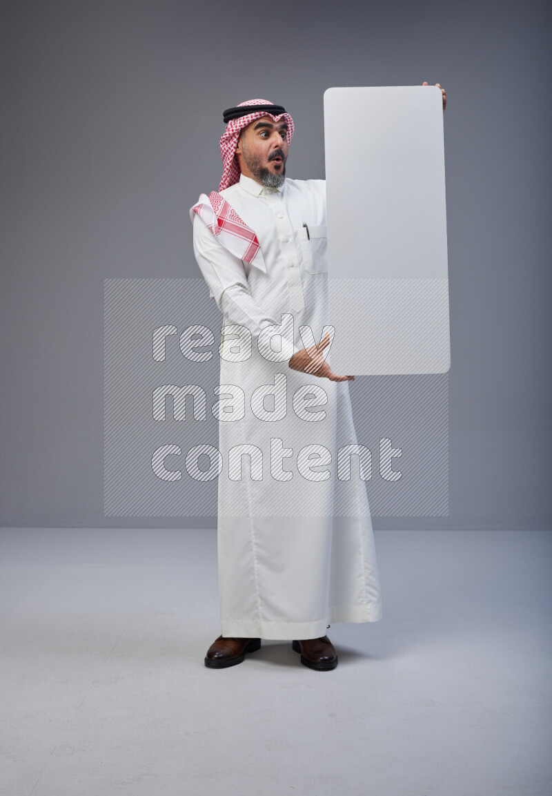 Saudi man Wearing Thob and red Shomag standing holding board on Gray background