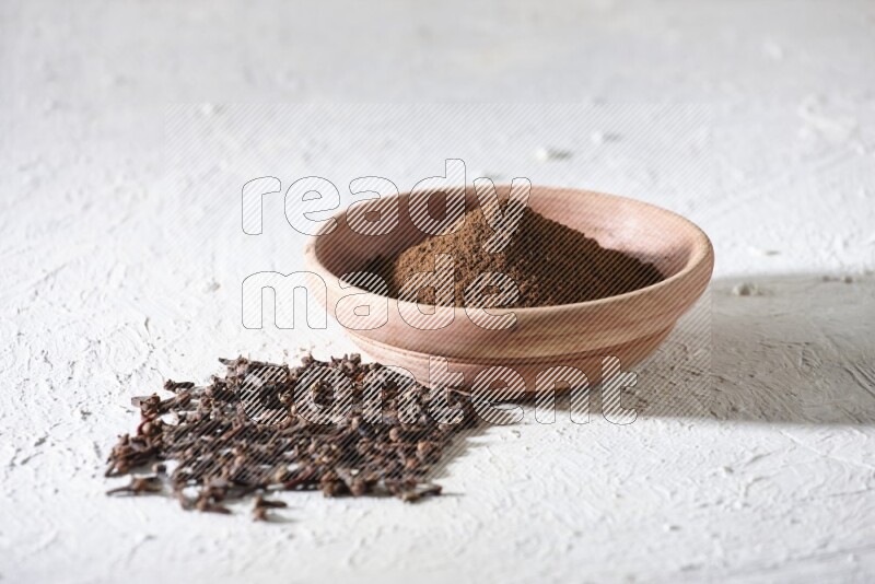 A wooden bowl full of cloves powder with whole cloves beside it on a textured white flooring