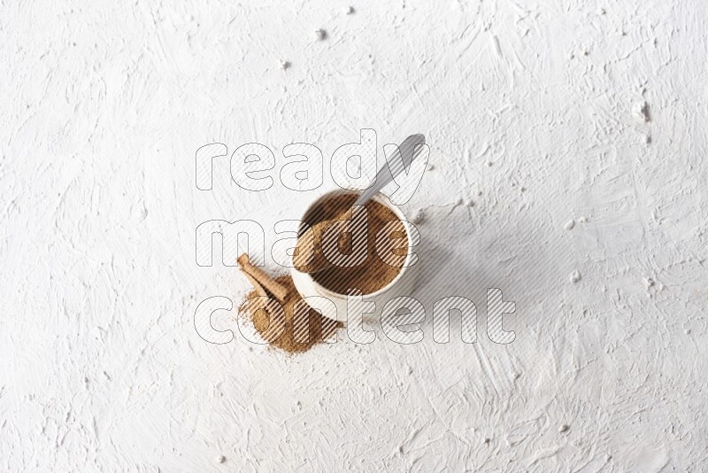 Ceramic beige bowl full of cinnamon powder and a metal spoon with cinnamon sticks next of it on a textured white background