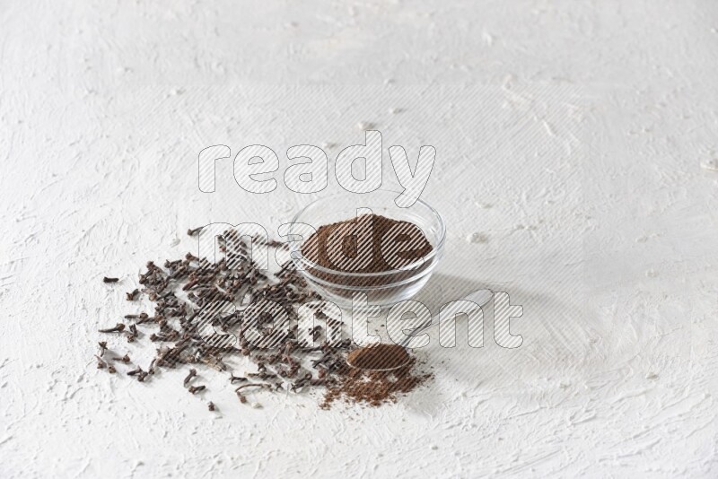 A glass bowl and a metal spoon full of cloves powder with cloves grains spread on a textured white flooring
