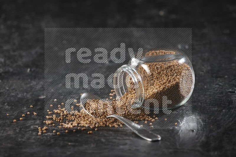 A glass spice jar and a metal spoon full of mustard seeds and jar is flipped with fallen seeds on a textured black flooring