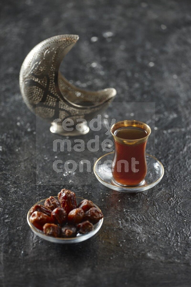 A silver lantern with different drinks, dates, nuts, prayer beads and quran on textured black background
