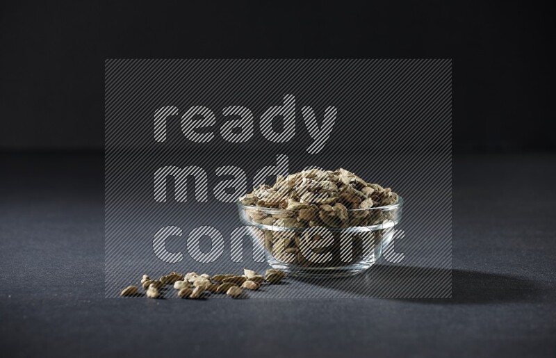 A glass bowl full of cardamom and more seeds spreaded beneath the bowl on black flooring
