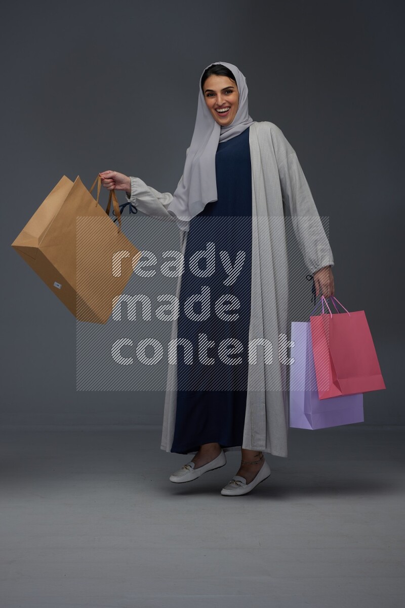 A Saudi woman wearing a light gray Abaya and head scarf standing and holding shopping bags on a grey background