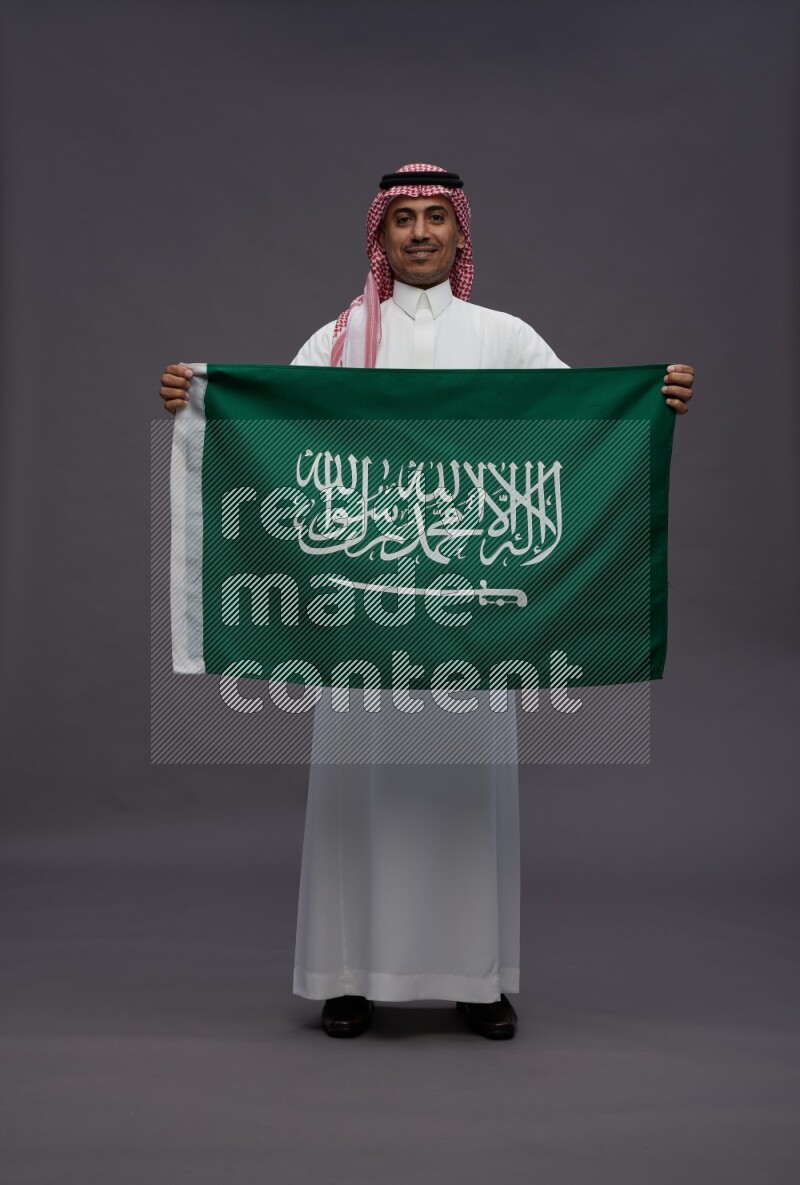 Saudi man wearing thob and shomag standing holding Saudi flag on gray background