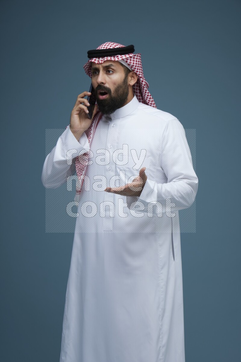 A man having a Call in a blue background wearing Saudi Thob with Shomag