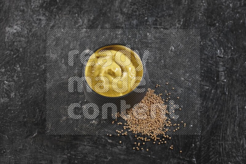 A black pottery bowl full of mustard paste with mustard seeds underneath on textured black flooring