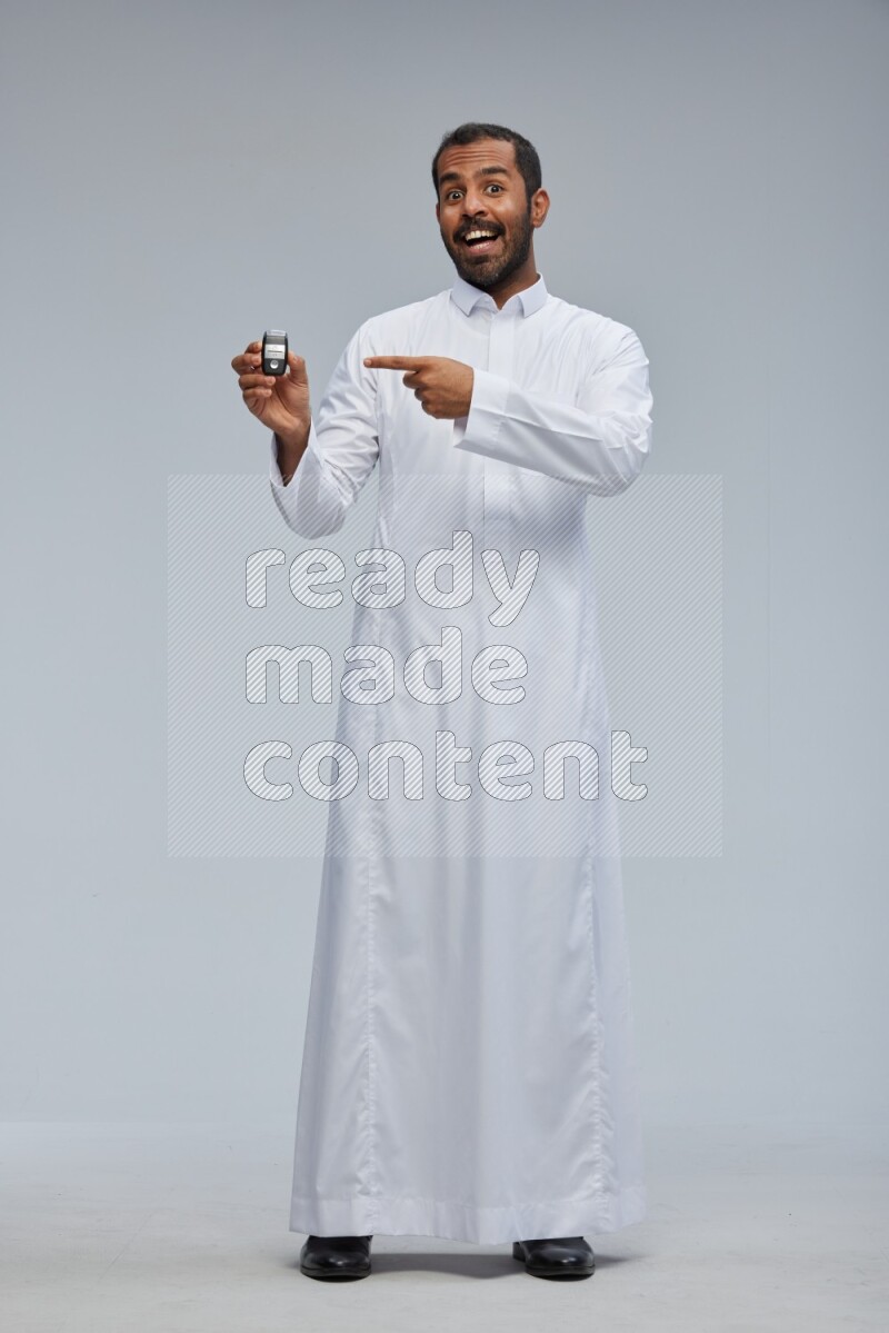 Saudi man wearing thob and shomag standing holding car key on gray background