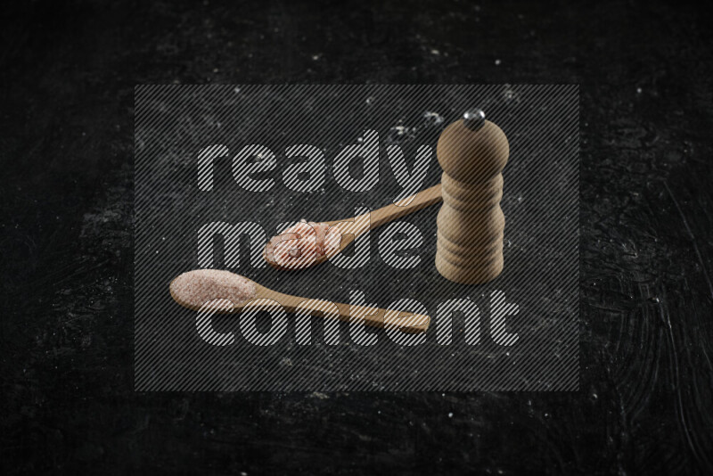 ملعقتان خشبيتان ممتلئتان بالملح الناعم والخشن وبجانبهما مطحنة خشبية على خلفية سوداء