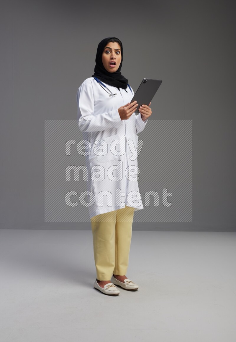 Saudi woman wearing lab coat with stethoscope standing working on tablet on Gray background