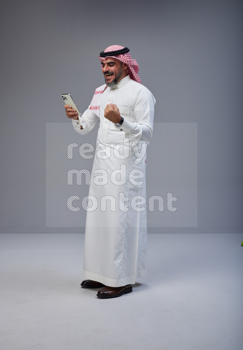 Saudi man Wearing Thob and red Shomag standing texting on phone on Gray background
