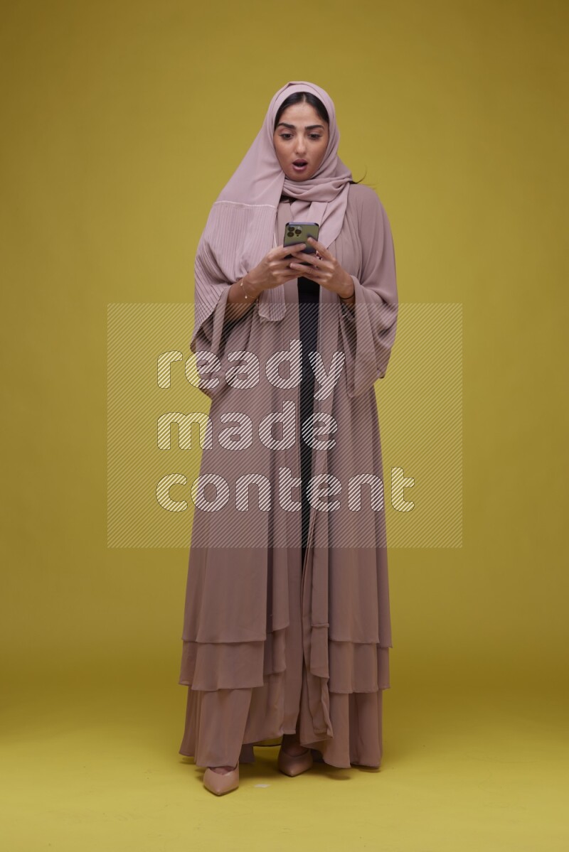 A woman Texting on her phone on a Yellow Background wearing Brown Abaya with Hijab