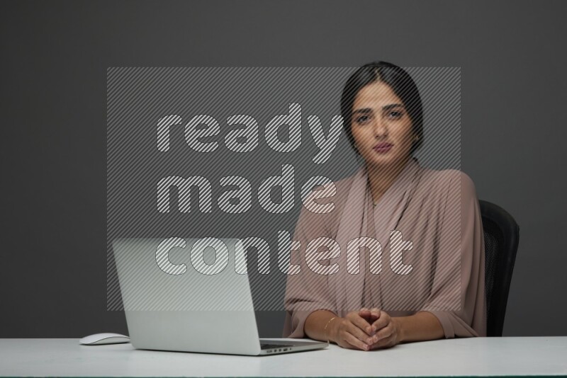 A Saudi woman Sitting on her desk on a Gray Background wearing Brown Abaya