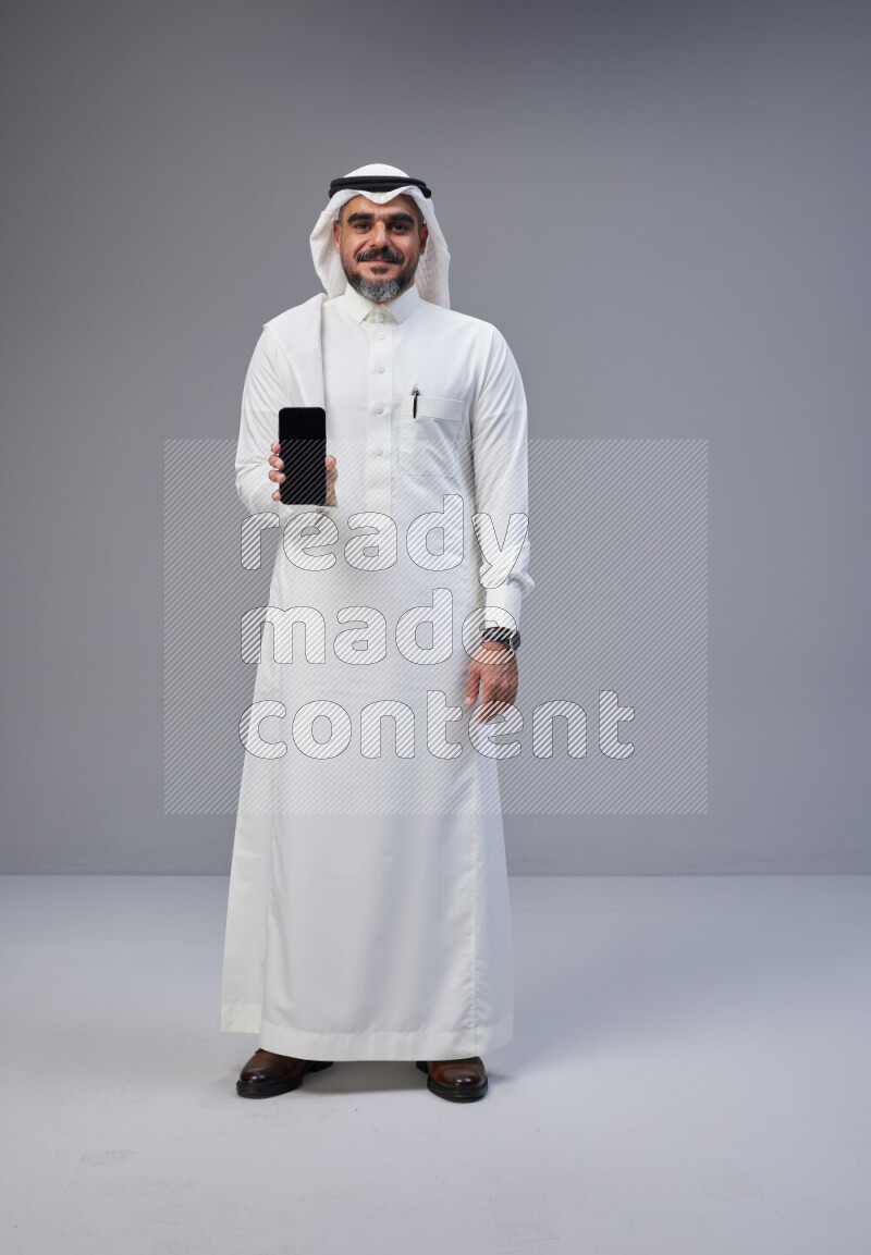 Saudi man Wearing Thob and white Shomag standing showing phone to camera on Gray background
