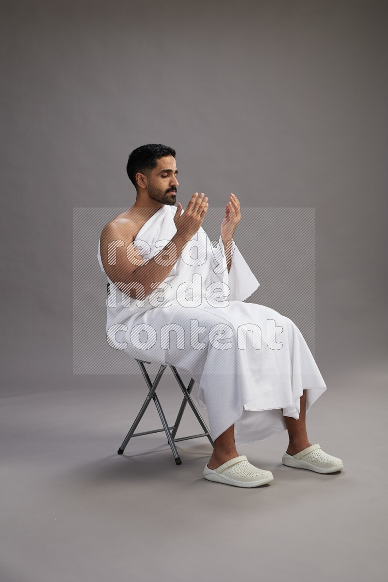 A man wearing Ehram sitting on chair performing dua'a on gray background