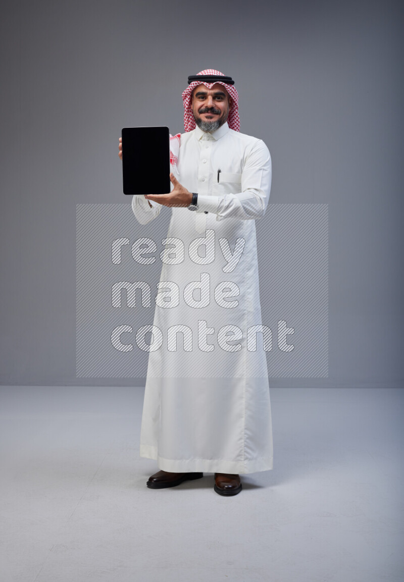 Saudi man Wearing Thob and red Shomag standing showing tablet to camera on Gray background