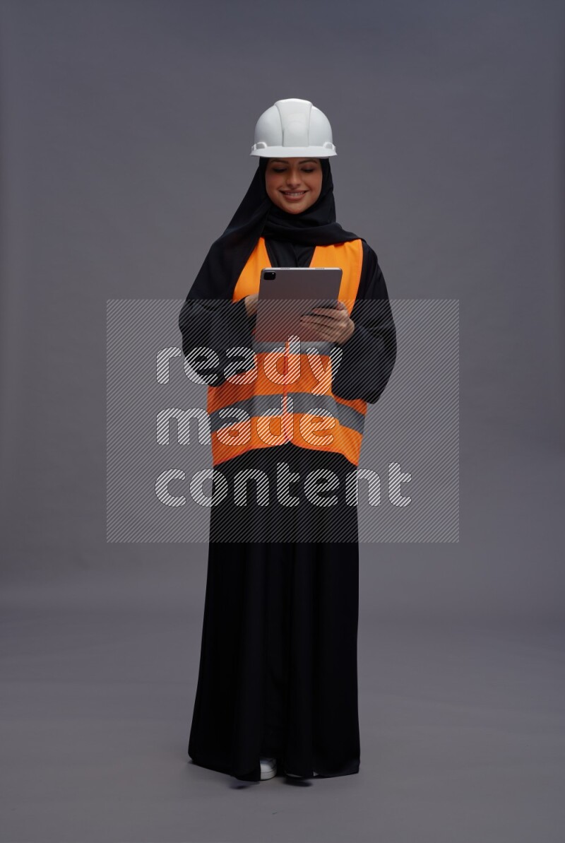 Saudi woman wearing Abaya with engineer vest standing working on tablet on gray background