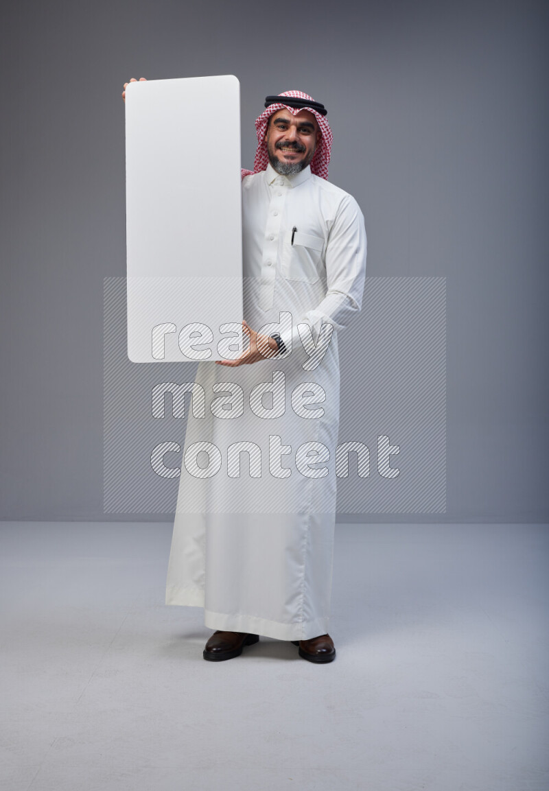 Saudi man Wearing Thob and red Shomag standing holding board on Gray background