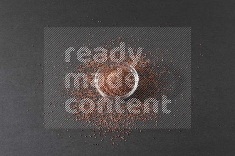 A glass bowl full of garden cress seeds surrounded by seeds on a black flooring