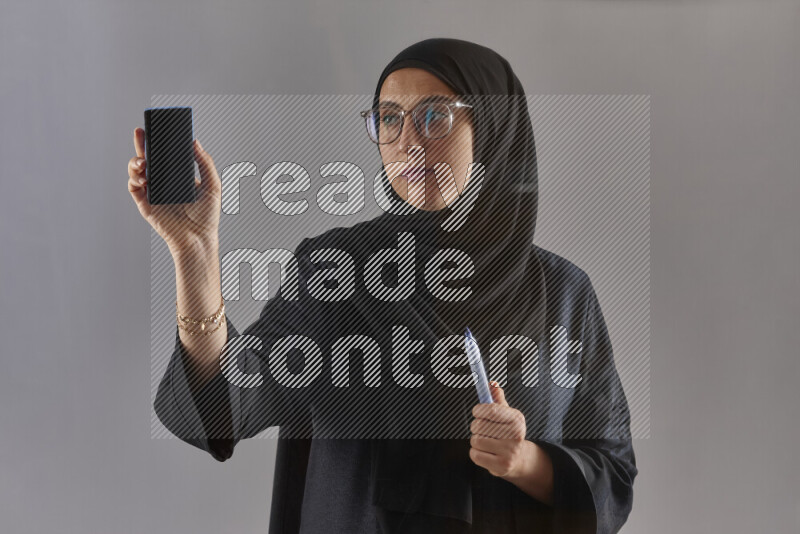 Woman in black abaya, hijab and glasses holding a marker pen to write on a transparent board with different reactions and expressions on grey background